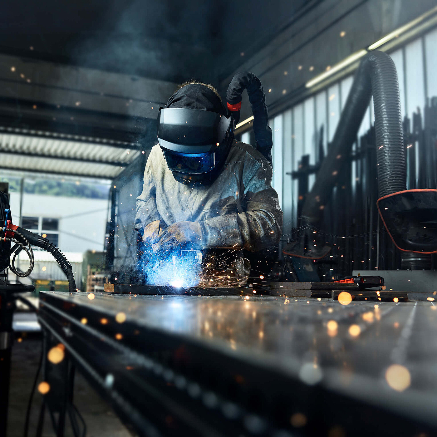 Schweißer mit Schutzmaske arbeitet an einem Metallstück in einer Werkstatt, Schweißgerät und Funken im Vordergrund.