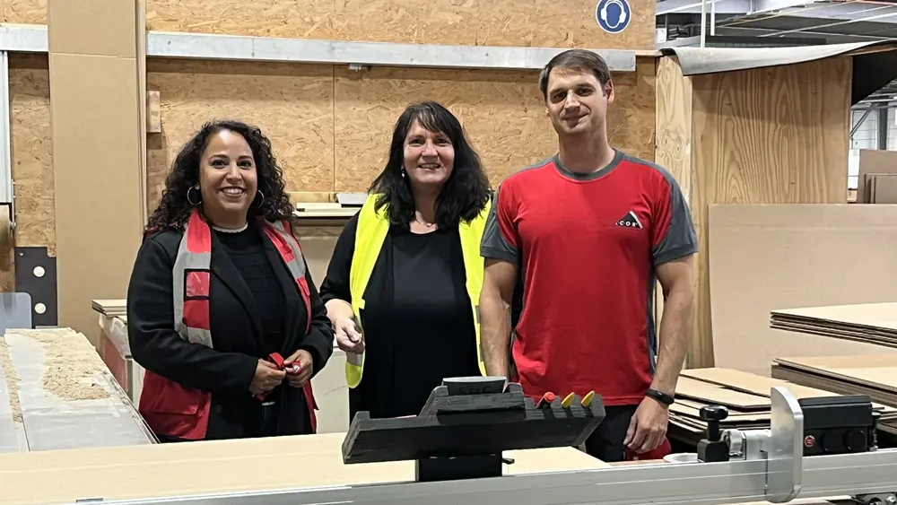 Drei Personen stehen lächelnd in einer Werkhalle vor einer Maschine; zwei tragen Arbeitskleidung, eine trägt eine gelbe Sicherheitsweste