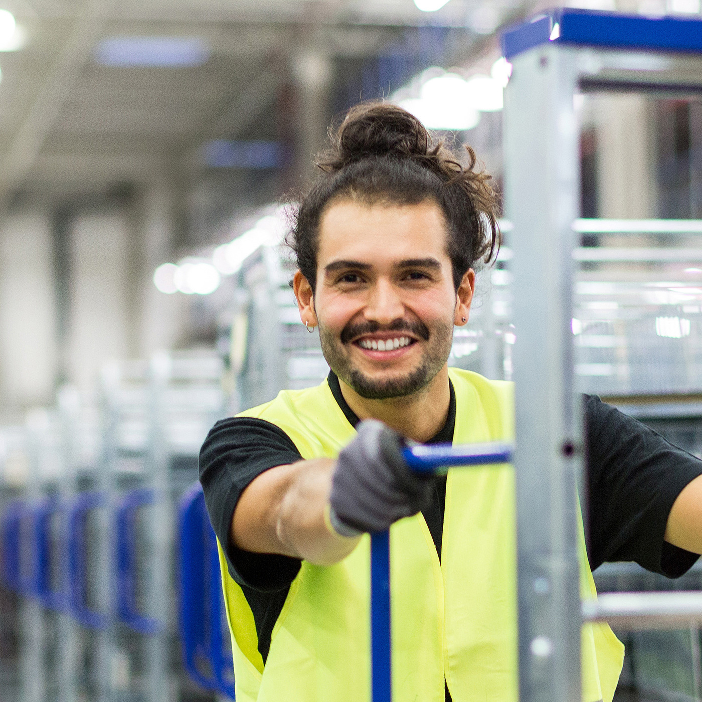 Produktionshelfer in einer Produktionshalle trägt eine gelbe Weste und bewegt einen Transportwagen.
